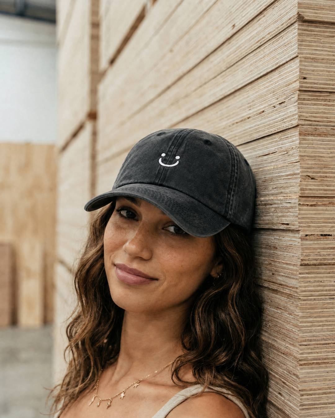 Woman wearing a black cap with a smiley face emblem against a wooden wall.