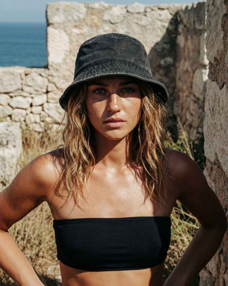Woman wearing a black bucket hat and black top standing against a stone wall with ocean view.