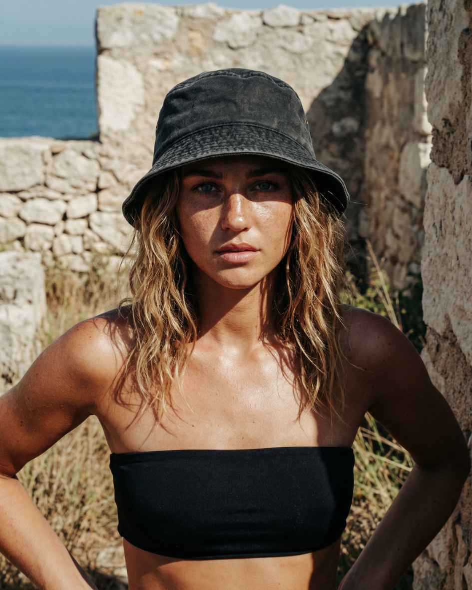 Woman wearing a black bucket hat and black top standing against a stone wall with ocean view.