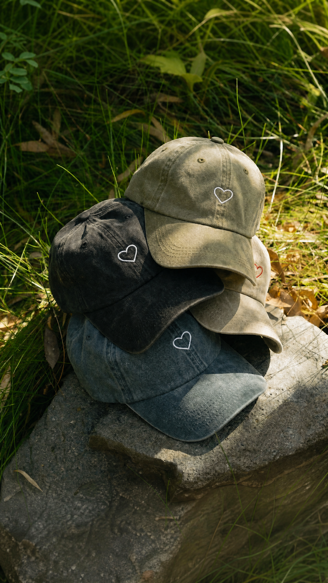 Three baseball caps with heart patterns on a stone surface surrounded by grass.