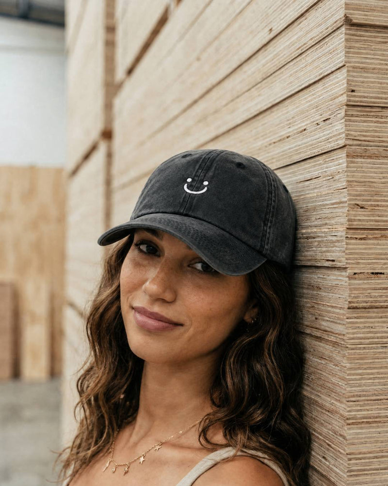 Woman wearing a black cap with a smiley face emblem against a wooden wall.