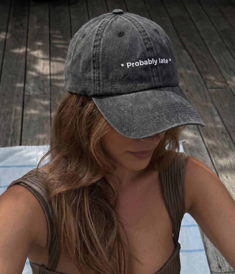 Person wearing a gray cap with 'Probably late' text on a wooden deck.